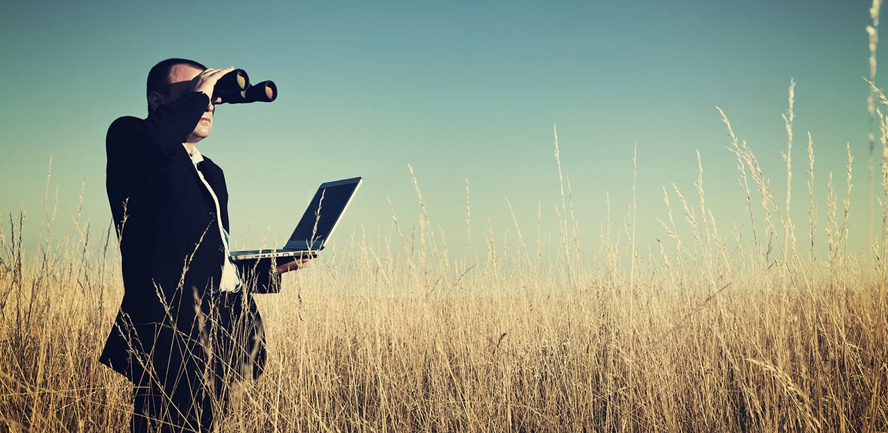Businessman in a field holding an open laptop and binoculars
